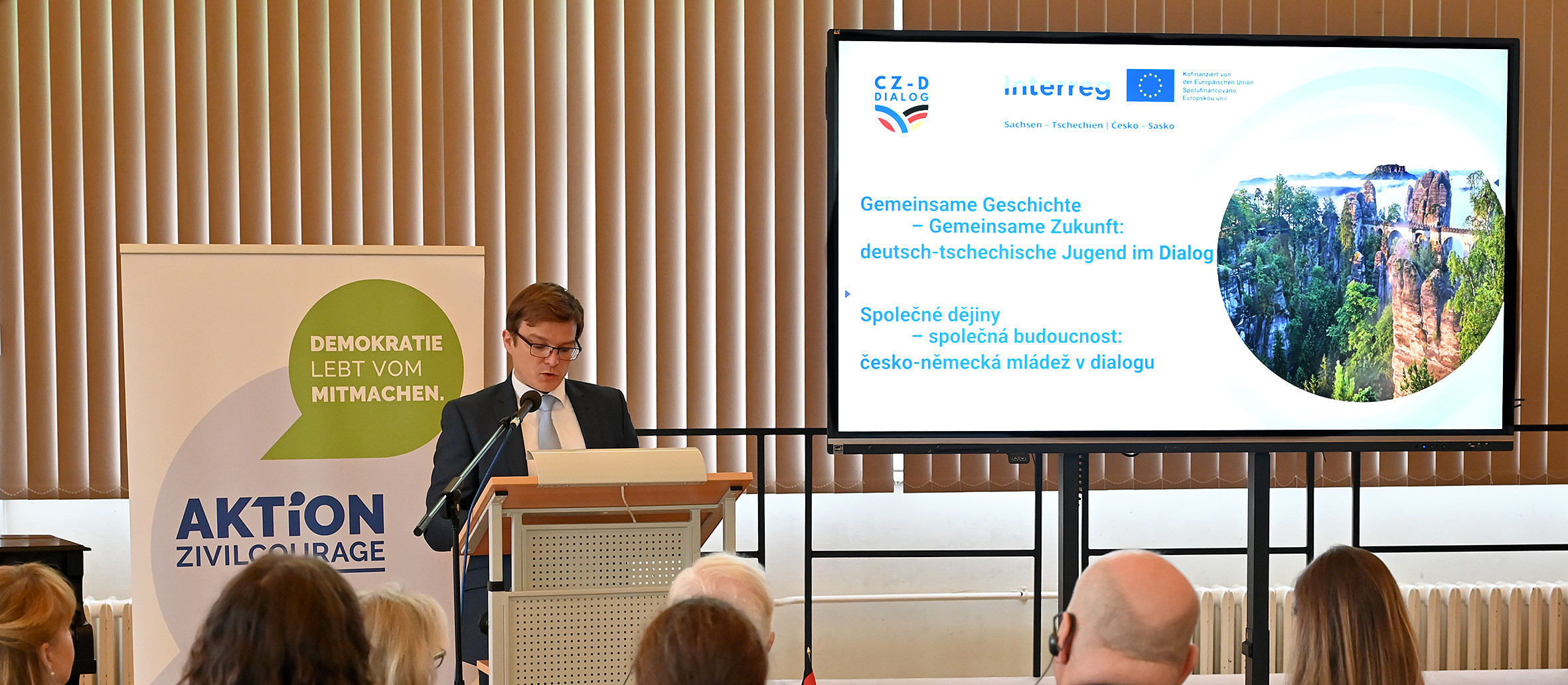 KI generiert: Das Bild zeigt einen Mann, der an einem Pult steht und eine Rede vor einem Publikum hält. Im Hintergrund sind eine Präsentation mit dem Titel "Gemeinsame Geschichte – Gemeinsame Zukunft: deutsch-tschechische Jugend im Dialog" und ein Banner mit der Aufschrift "AKTION Zivilcourage" zu sehen.