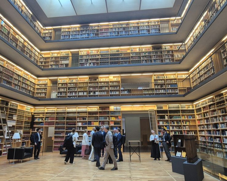 KI generiert: Das Bild zeigt eine formelle Besprechung oder Konferenz in einem historischen Raum mit großen Fenstern und verzierten Wänden. Mehrere Personen sitzen an langen Tischen, die in einem U-förmigen Arrangement angeordnet sind, und scheinen sich auf eine Diskussion oder Präsentation zu konzentrieren.