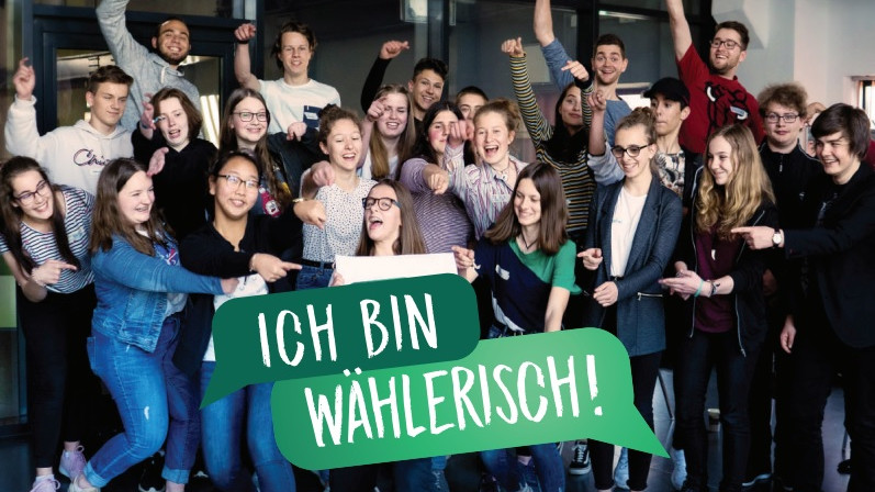 KI generiert: Das Bild zeigt eine Gruppe junger Menschen, die fröhlich und enthusiastisch posieren, während sie ein Schild mit der Aufschrift "ICH BIN WÄHLERISCH!" halten. Der Hauptinhalt des Bildes ist die Darstellung von Jugendlichen, die ihr Interesse oder ihre Bereitschaft zur Wahlbeteiligung ausdrücken.