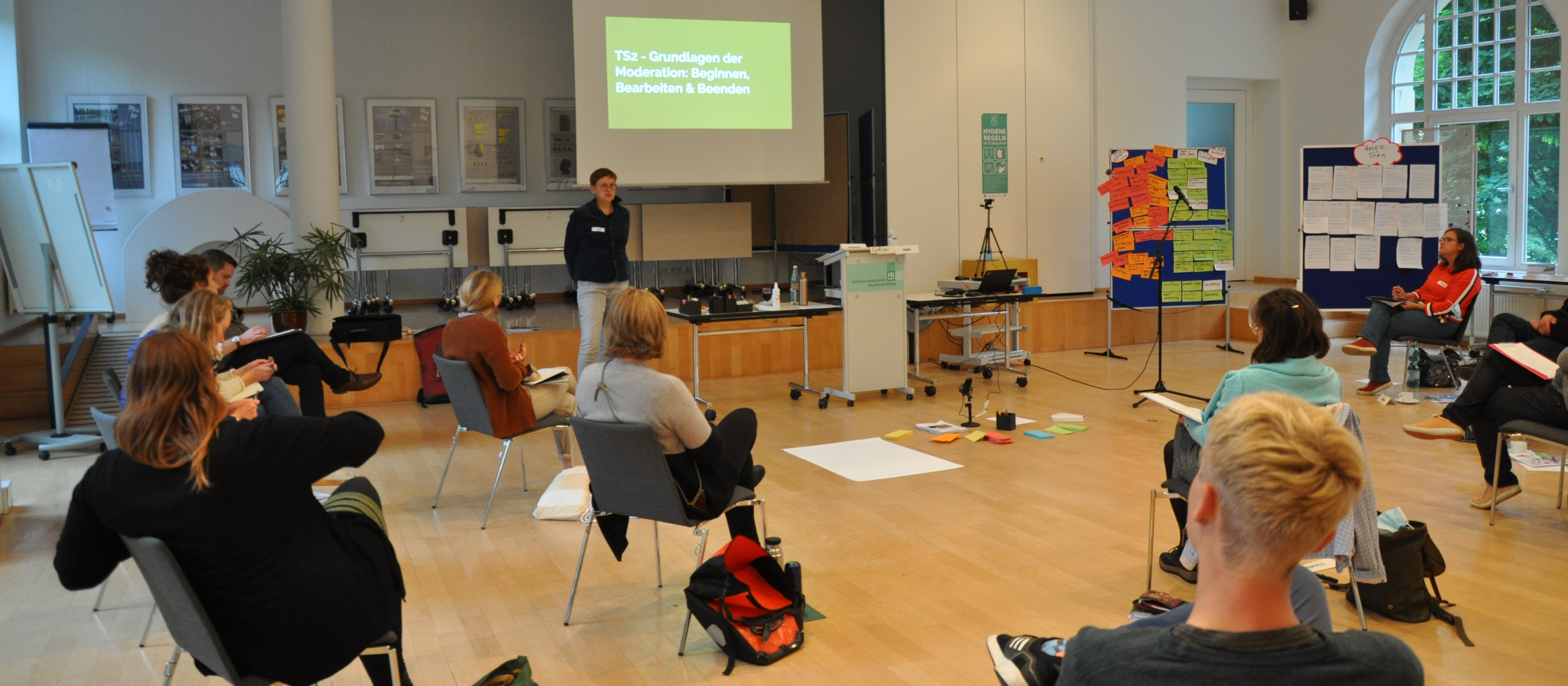 KI generiert: Das Bild zeigt eine Gruppe von Personen in einem Seminarraum, die an einer Präsentation oder einem Workshop teilnehmen. Der Hauptinhalt ist eine Bildungsveranstaltung, bei der ein Referent vorne steht und eine Präsentation hält, während die Teilnehmer zuhören und Notizen machen.
