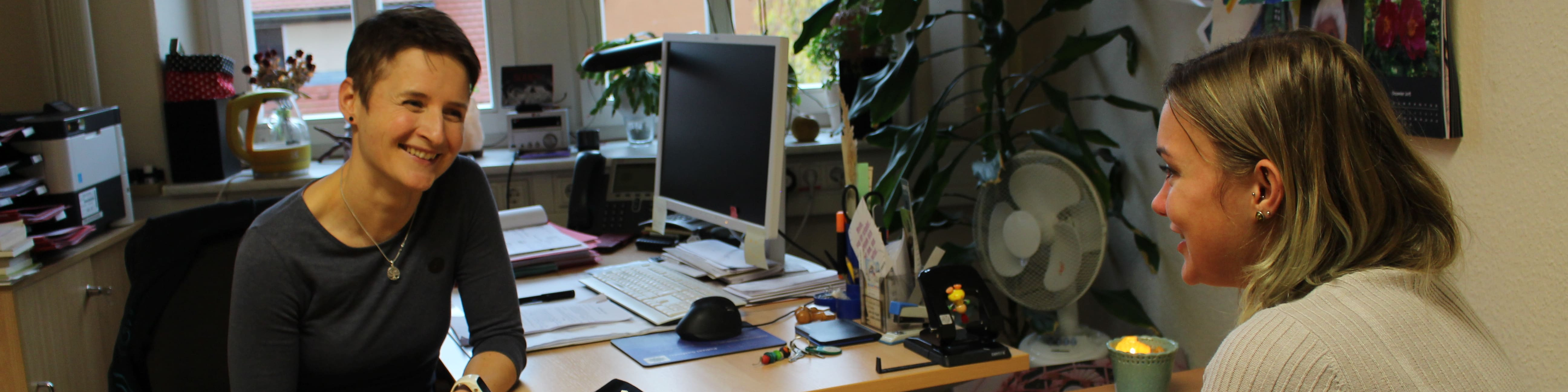 KI generiert: Das Bild zeigt zwei Personen in einem Büro, wobei eine Person an einem Schreibtisch sitzt und die andere Person steht und mit ihr spricht. Der Hauptinhalt des Bildes ist eine Büroumgebung mit Computern, Pflanzen und verschiedenen Büromaterialien.