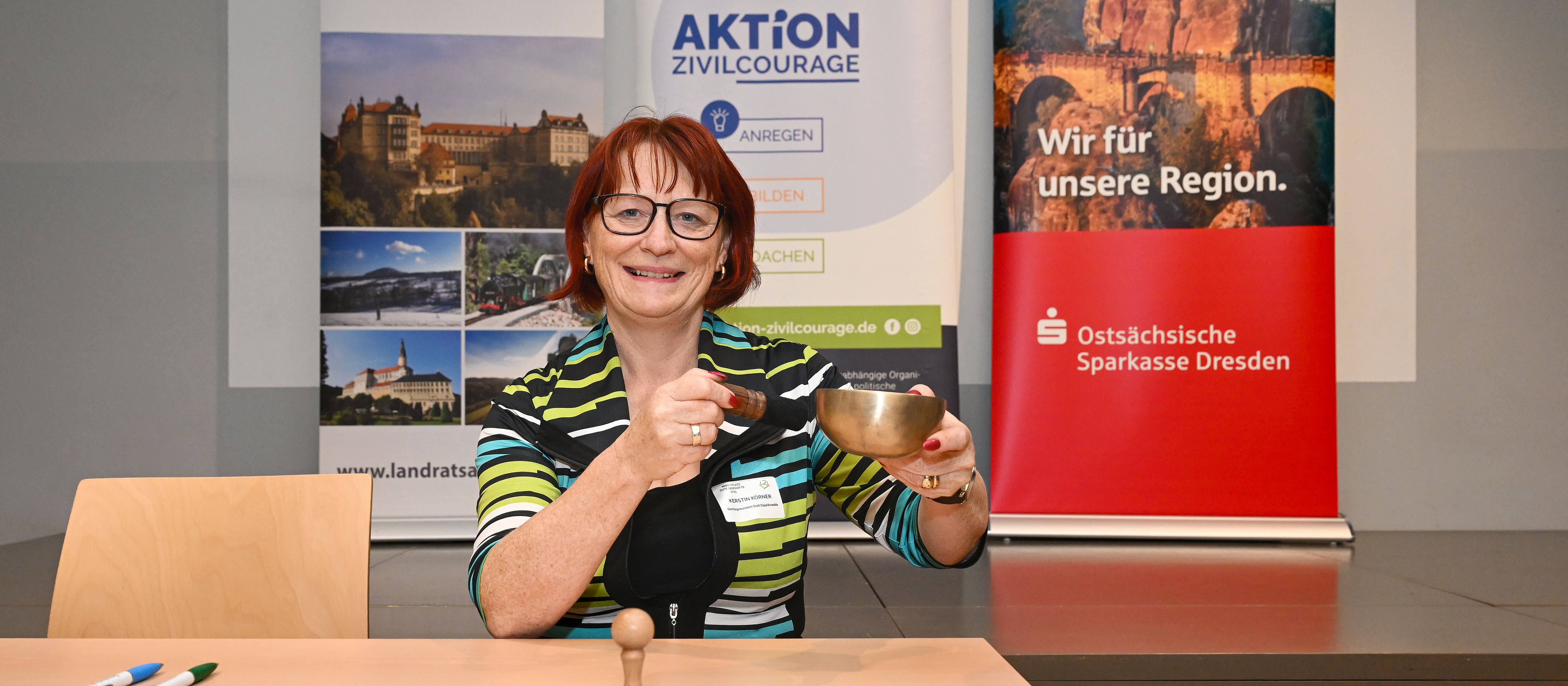 KI generiert: Das Bild zeigt eine Frau, die lächelnd an einem Tisch sitzt und eine Glocke in der Hand hält. Im Hintergrund sind zwei Roll-up-Banner mit Informationen über die Region Dresden und die Aktion Zivilcourage zu sehen.