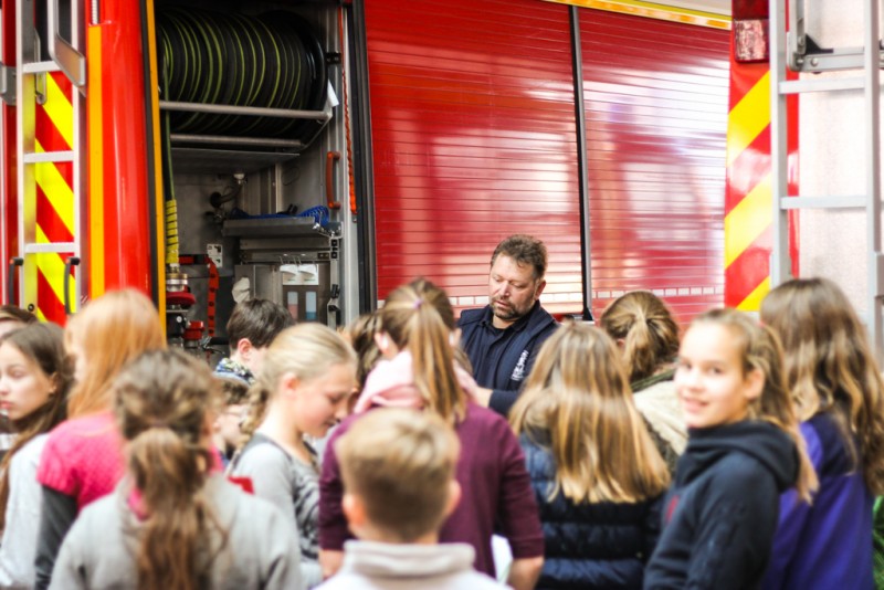 KI generiert: Das Bild zeigt eine Gruppe von Kindern und Jugendlichen, die sich um einen Mann in Uniform versammelt haben, der vor einem roten Feuerwehrfahrzeug steht. Der Hauptinhalt des Bildes ist eine Bildungsveranstaltung oder Führung, bei der die Kinder etwas über die Feuerwehr und deren Ausrüstung lernen.