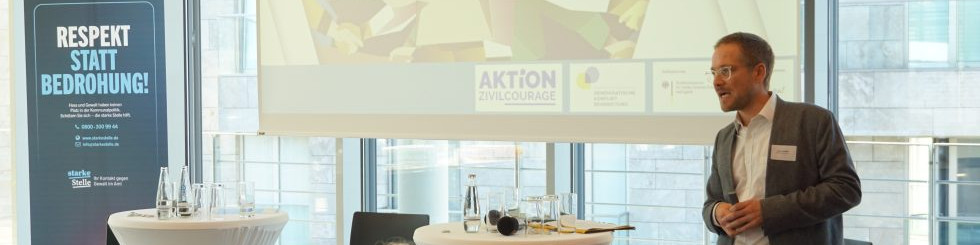 KI generiert: Das Bild zeigt einen Mann, der in einem Konferenzraum vor einem Publikum spricht. Im Hintergrund sind ein Banner mit der Aufschrift "AKTION ZIVILCOURAGE" und ein weiteres Banner mit der Aufschrift "RESPEKT STATT BEDROHUNG!" sichtbar.