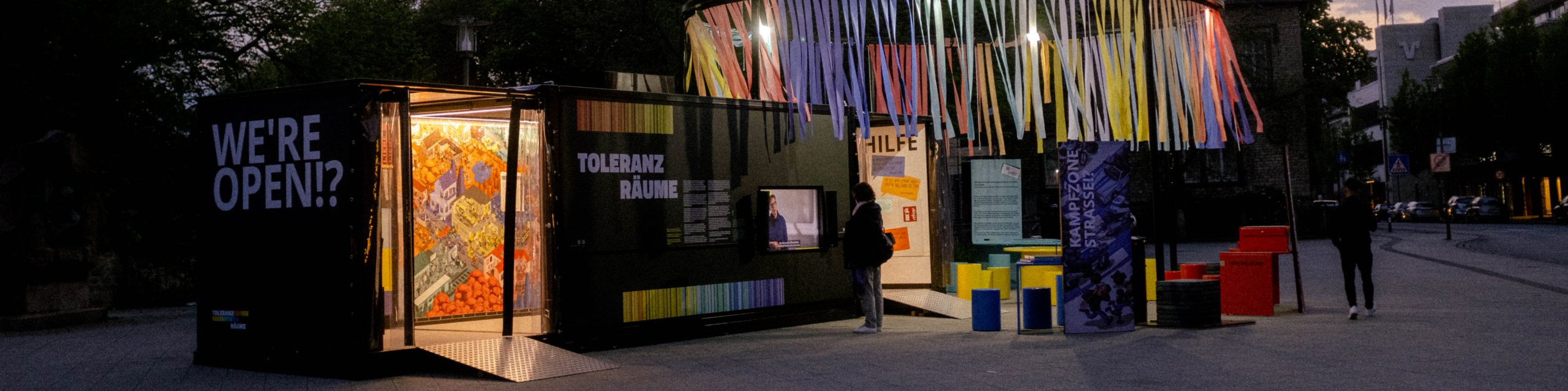 KI generiert: Das Bild zeigt eine nächtliche Szene mit einem beleuchteten Container, der als Ausstellungsraum genutzt wird und auf dem "WE'RE OPEN!" steht. Im Hintergrund sind bunte Bänder und weitere Ausstellungsstände zu sehen, die auf eine kulturelle oder künstlerische Veranstaltung hinweisen.