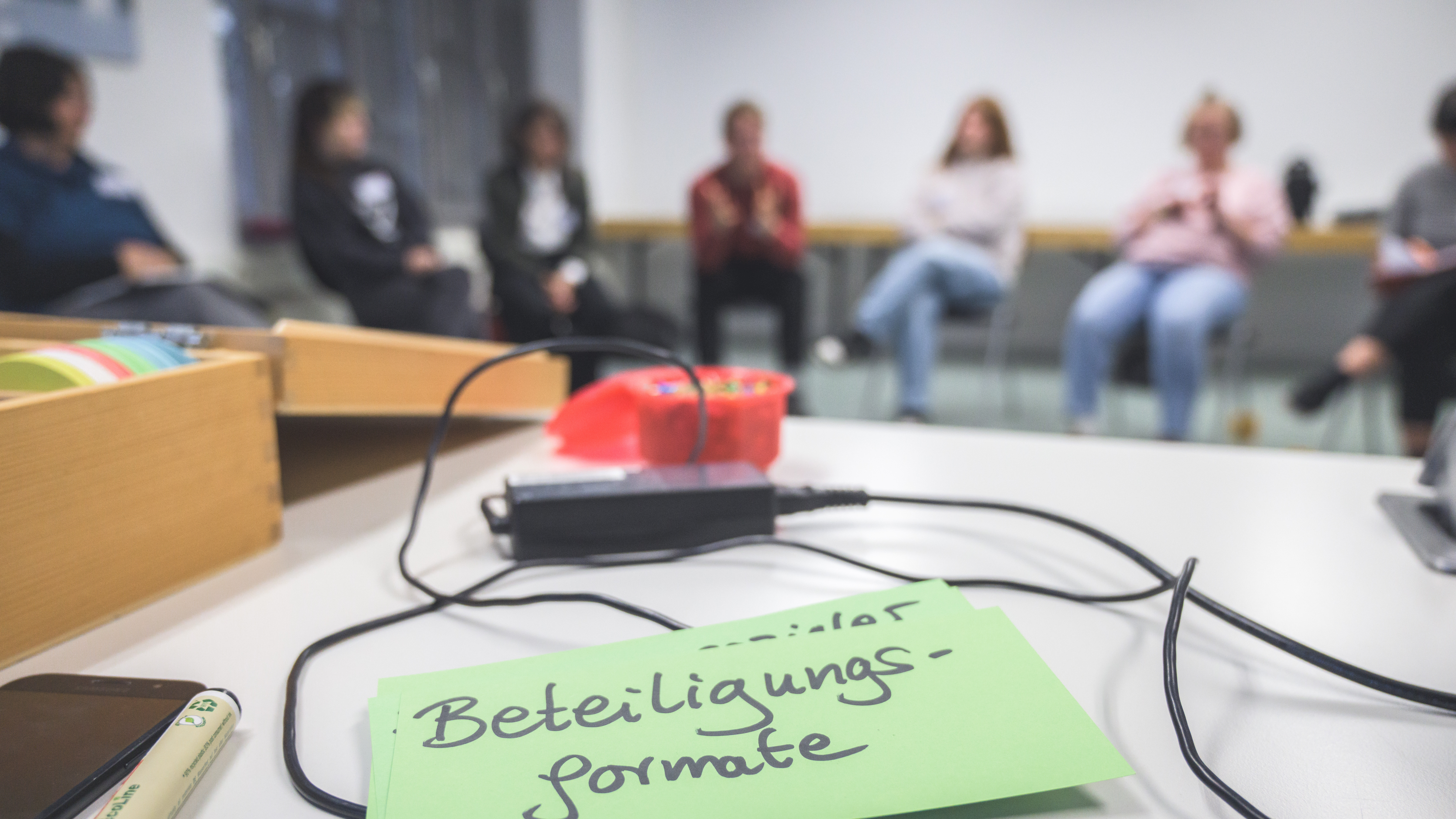 KI generiert: Das Bild zeigt eine Gruppe von Personen, die in einem Raum sitzen und sich unterhalten, während im Vordergrund ein grünes Blatt mit der Aufschrift "Beteiligungsformate" liegt, daneben ein rotes Mikrofon und einige Kabel. Der Hauptinhalt des Bildes scheint eine Diskussion oder ein Workshop zu Partizipationsmethoden zu sein.