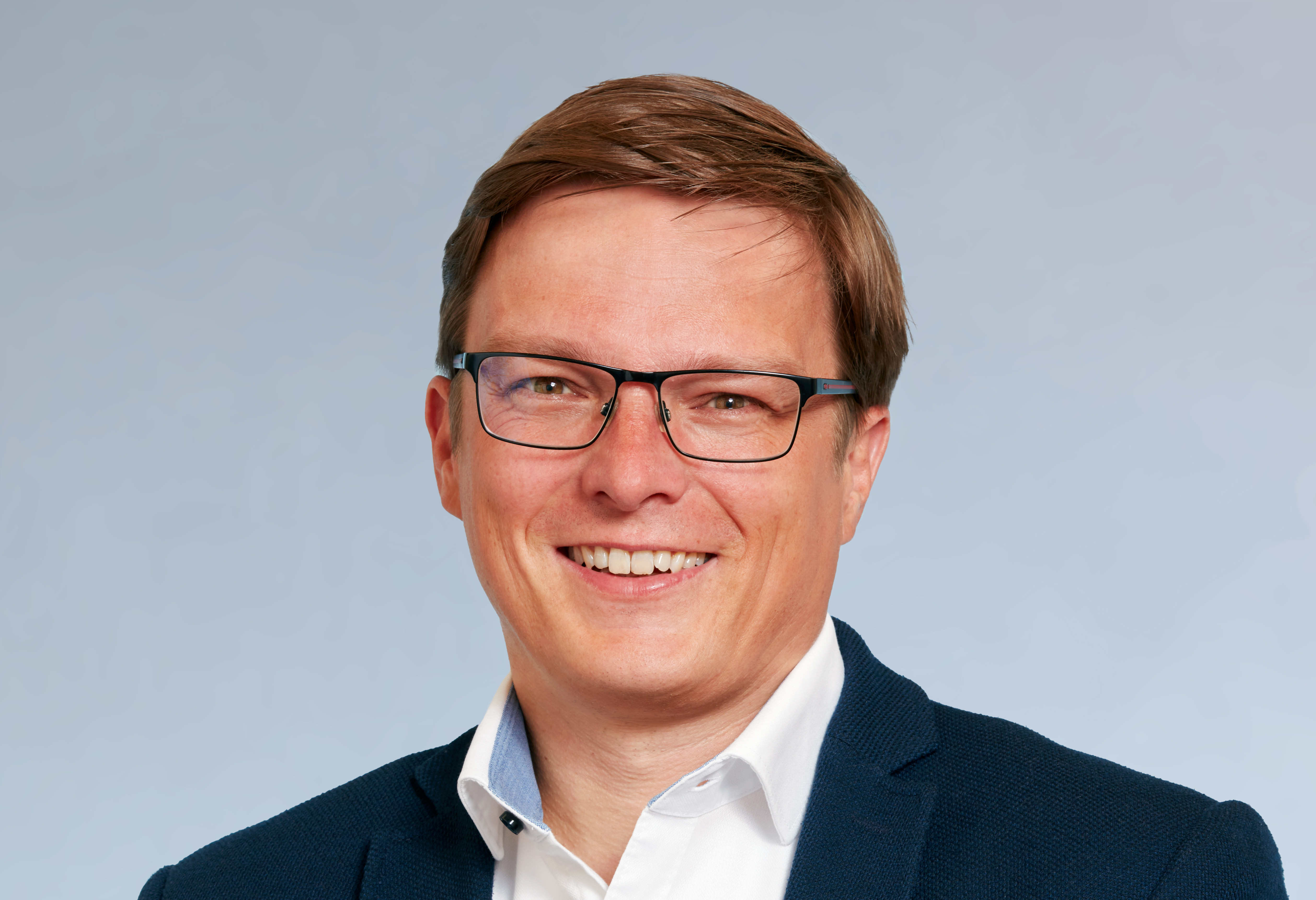 KI generiert: Das Bild zeigt einen lächelnden Mann mit kurzen braunen Haaren und einer Brille. Er trägt ein weißes Hemd mit einem dunklen Blazer und hat einen neutralen Hintergrund.