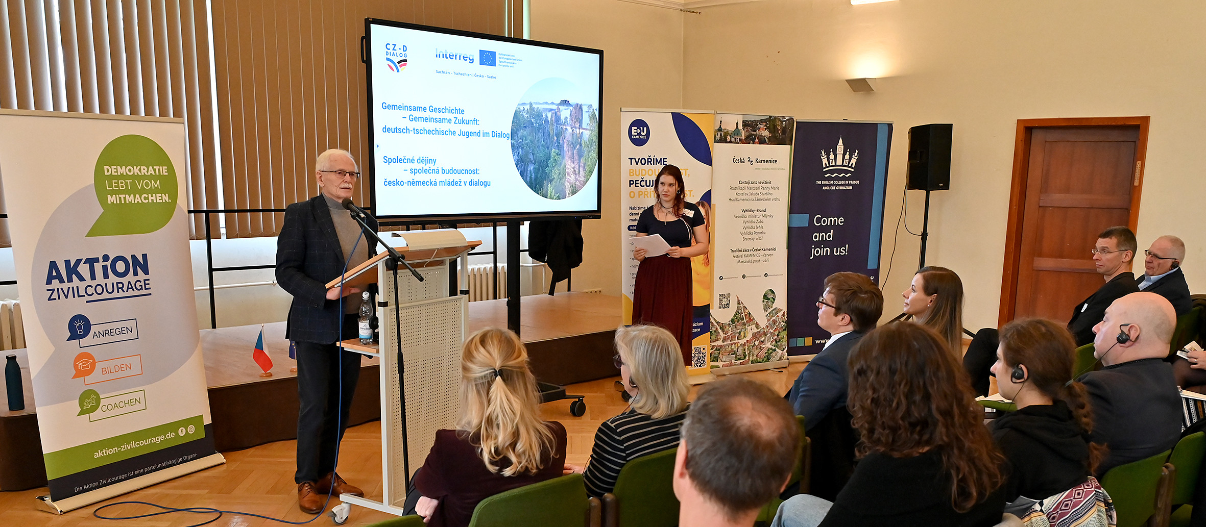 KI generiert: Das Bild zeigt eine Präsentation oder Diskussion in einem Konferenzraum, bei der ein Mann am Rednerpult spricht und eine Frau daneben steht. Im Hintergrund sind verschiedene Banner und eine Leinwand mit einer Präsentation zu sehen, und mehrere Personen sitzen im Publikum.