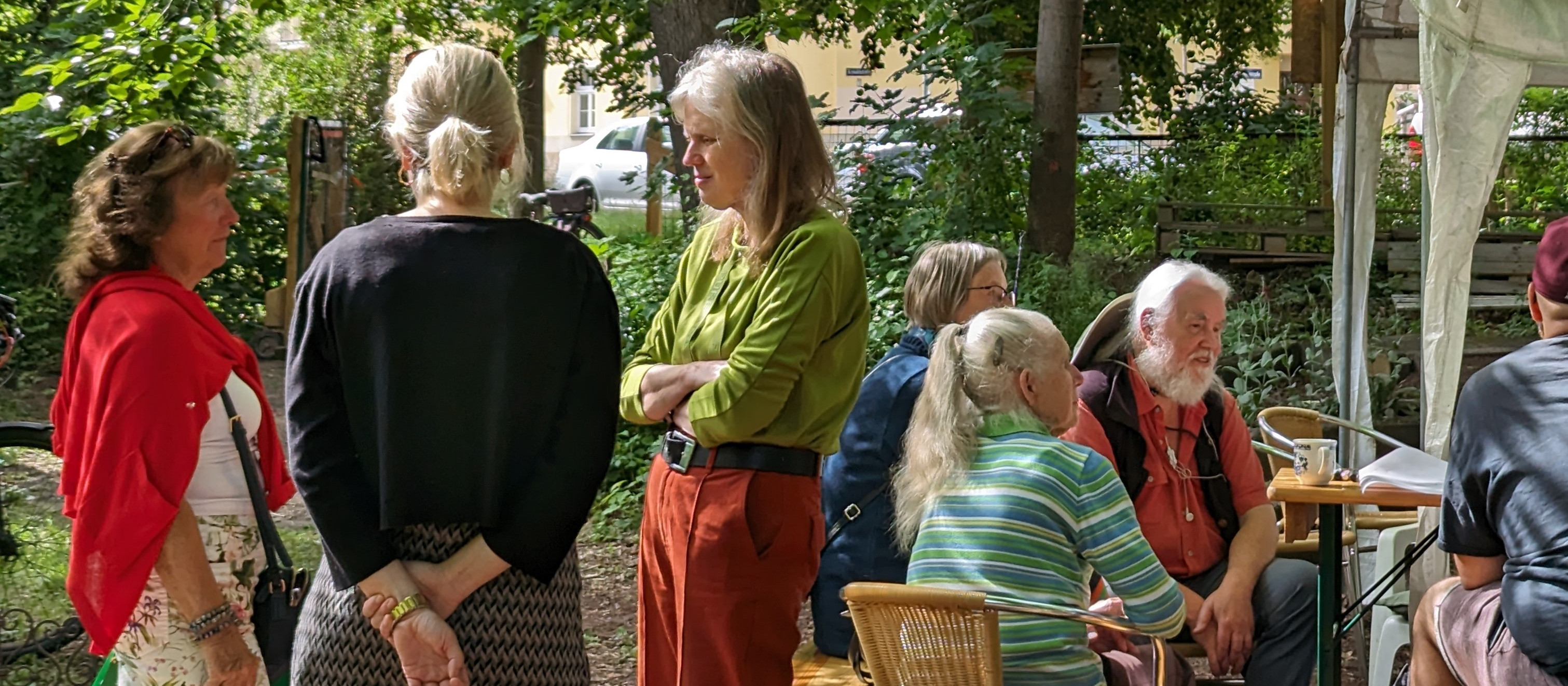 KI generiert: Das Bild zeigt eine Gruppe von Menschen, die sich in einem grünen, parkähnlichen Bereich versammelt haben, einige stehen und unterhalten sich, während andere auf Stühlen sitzen. Der Hauptinhalt des Bildes ist eine gesellige Zusammenkunft im Freien, möglicherweise ein Treffen oder eine kleine Veranstaltung.