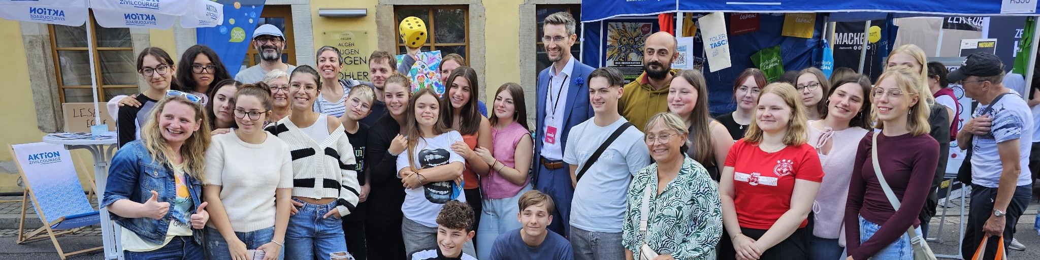 KI generiert: Das Bild zeigt eine Gruppe von Menschen, die vor einem Stand mit dem Schriftzug "AKTION" posieren. Es handelt sich vermutlich um eine Veranstaltung oder ein Event, bei dem die Teilnehmer gemeinsam ein Foto machen.