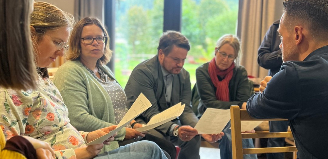 KI generiert: Das Bild zeigt eine Gruppe von fünf Personen, die in einem hellen Raum mit großen Fenstern sitzen und sich scheinbar über Dokumente oder Notizen austauschen. Der Hauptinhalt des Bildes ist eine Besprechung oder ein Workshop, bei dem die Teilnehmer aktiv diskutieren und zusammenarbeiten.