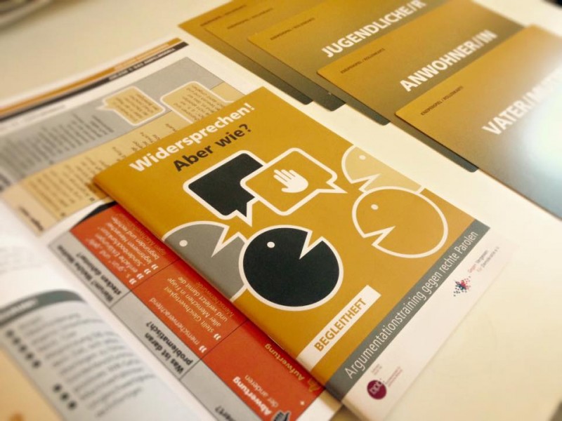 KI generiert: Das Bild zeigt mehrere Broschüren und Informationsblätter, die auf einem Tisch liegen. Der Hauptinhalt der sichtbaren Broschüre bezieht sich auf das Thema "Widersprechen! Aber wie?" und scheint sich mit Argumentationstraining gegen rechte Parolen zu befassen.