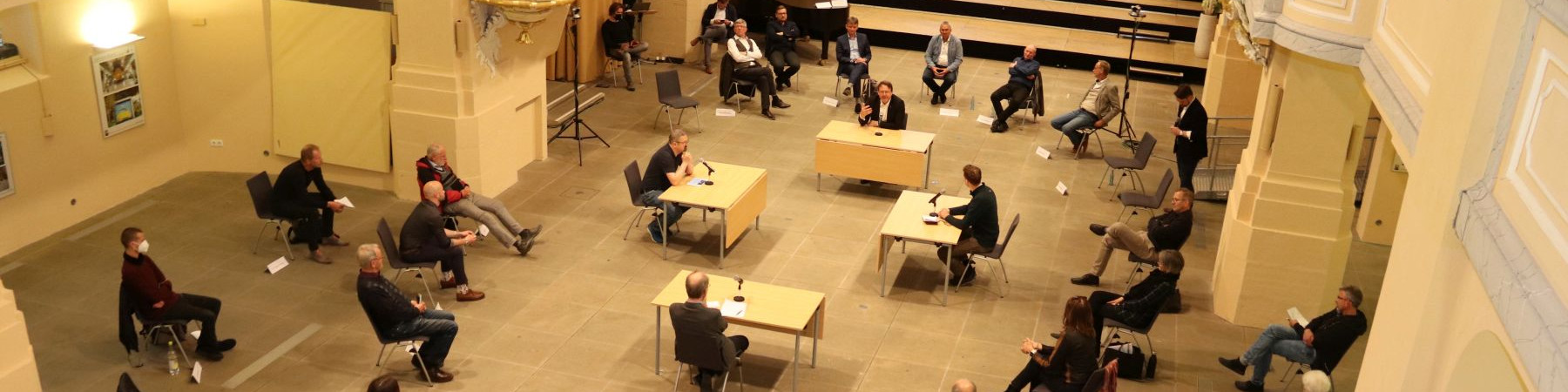 KI generiert: Das Bild zeigt eine Versammlung oder ein Meeting in einem großen, hellen Raum mit hohen Decken und Säulen. Im Mittelpunkt stehen mehrere Personen an Tischen mit Mikrofonen, während andere im Publikum sitzen und zuhören.