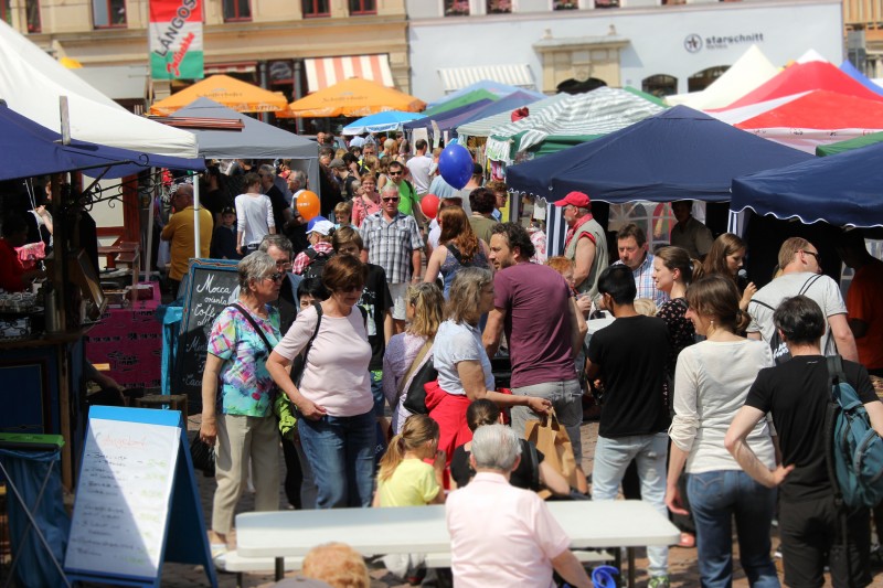 KI generiert: Das Bild zeigt eine belebte Straßenmarkt-Szene mit vielen Menschen, die zwischen bunten Marktständen mit verschiedenen Waren und Speisen umherlaufen. Der Hauptinhalt des Bildes ist der lebhafte Markt mit einer vielfältigen Menge an Besuchern und farbenfrohen Ständen.