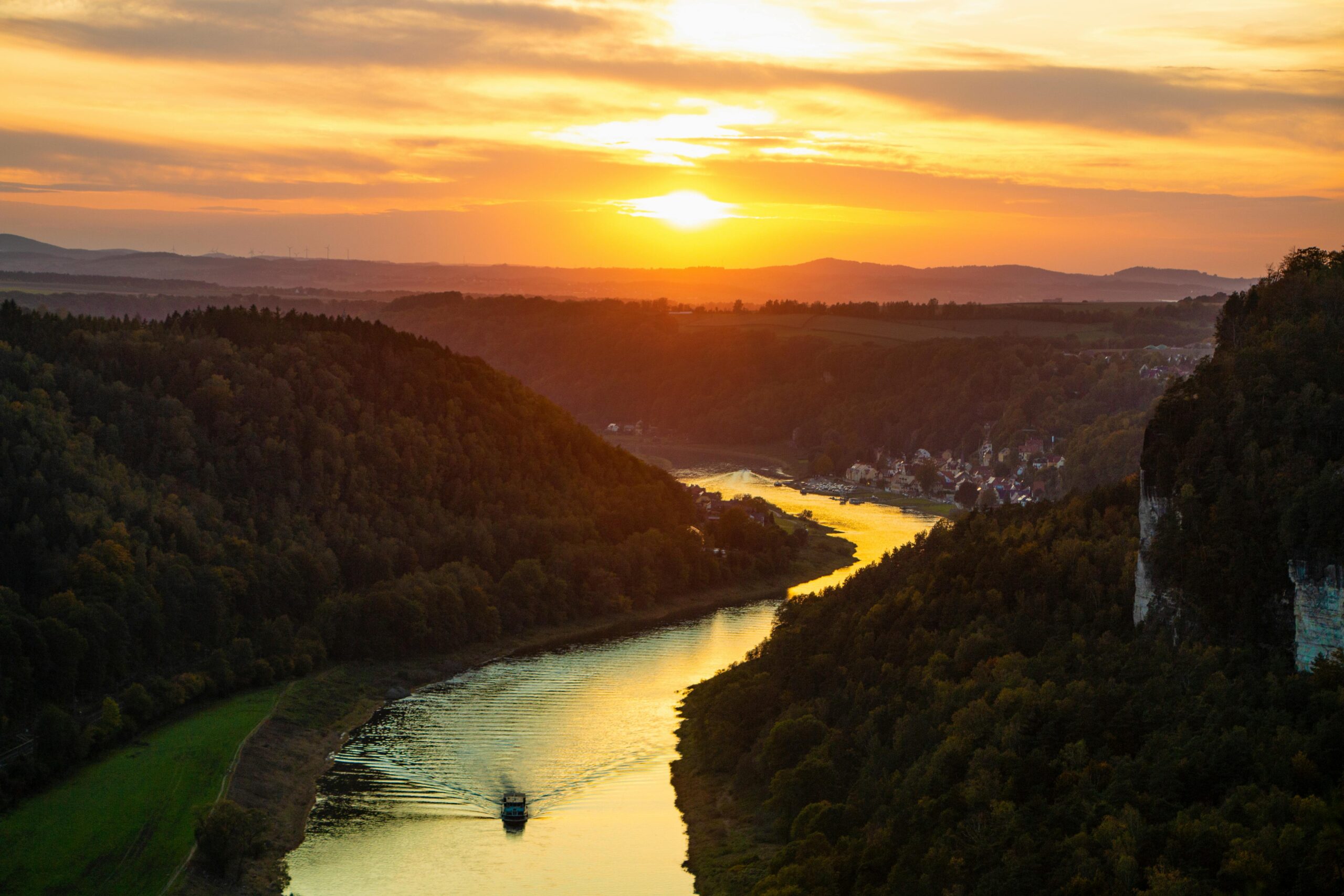 KI generiert: Das Bild zeigt eine malerische Flusslandschaft bei Sonnenuntergang, mit einem Boot auf dem Fluss, umgeben von bewaldeten Hügeln und einem kleinen Dorf in der Ferne. Der Hauptinhalt des Bildes ist die harmonische Natur mit dem Fluss, der Landschaft und dem Sonnenuntergang.