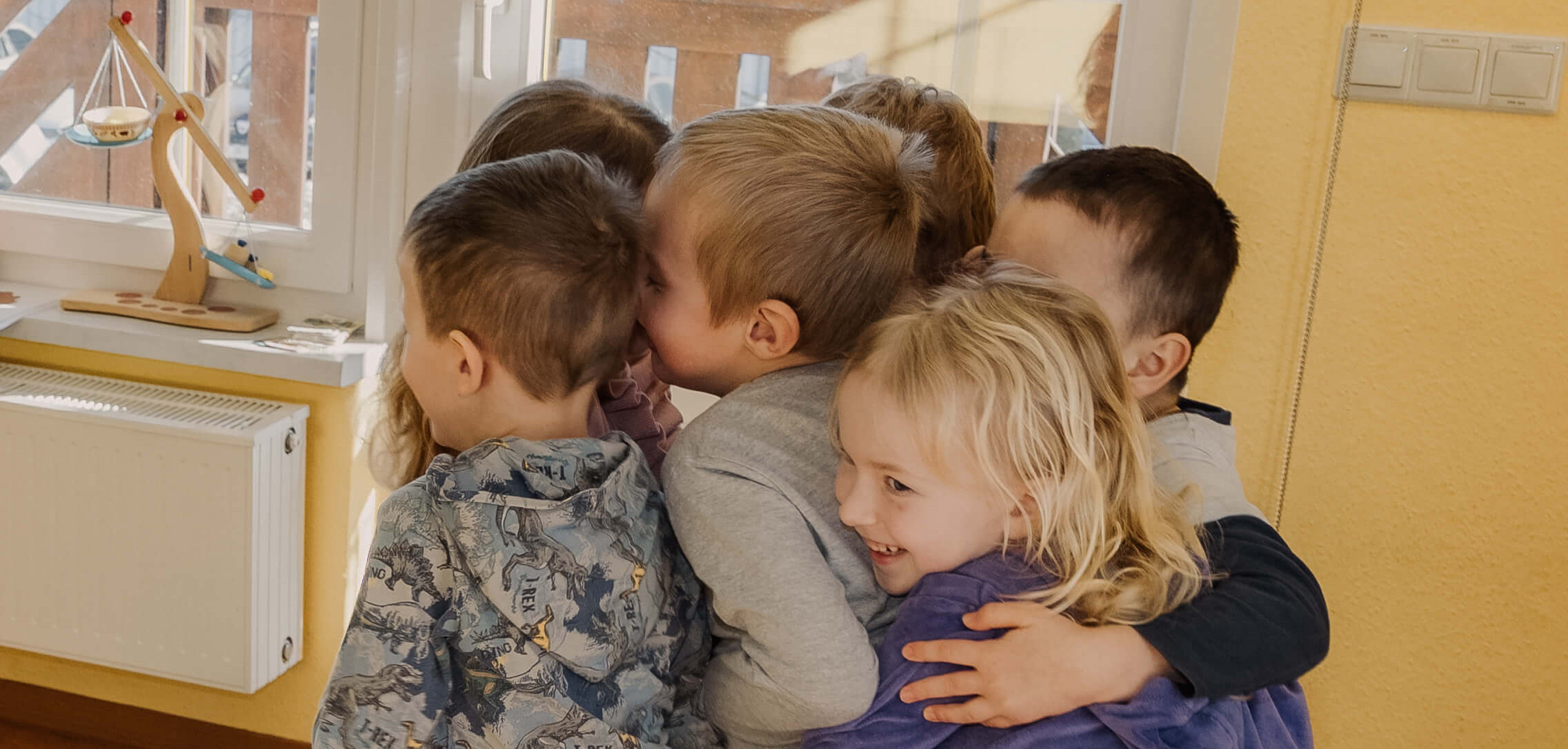KI generiert: Das Bild zeigt eine Gruppe von Kindern, die sich in einem Raum umarmen und gemeinsam lachen. Im Hintergrund sind ein Fenster und ein Regal mit einem Spielzeug zu sehen.