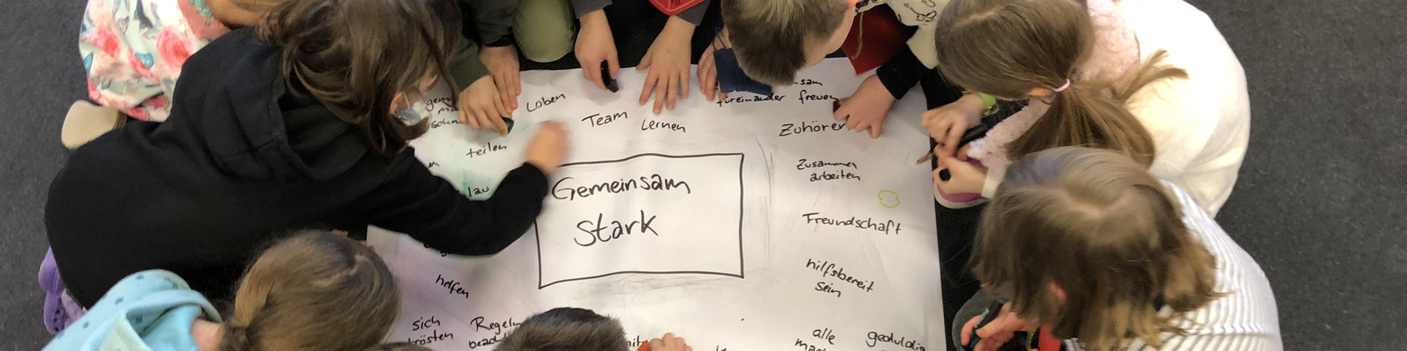 KI generiert: Das Bild zeigt eine Gruppe von Kindern, die gemeinsam auf einem großen Blatt Papier Ideen sammeln und aufschreiben. Der Hauptinhalt ist eine Brainstorming-Sitzung, bei der die Kinder das Thema "Gemeinsam Stark" bearbeiten.