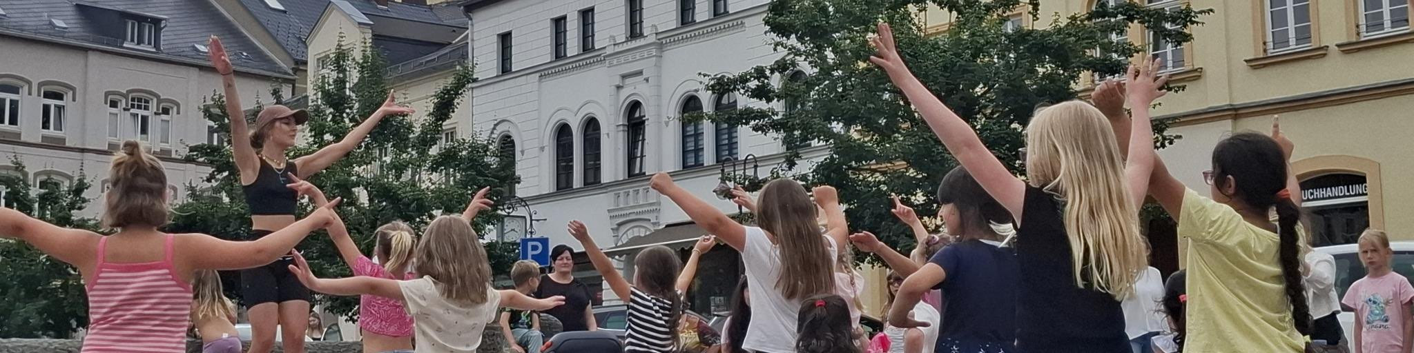 KI generiert: Das Bild zeigt eine Gruppe von Kindern und Jugendlichen, die draußen eine Tanz- oder Bewegungsaktivität durchführen, angeleitet von einer erwachsenen Person. Im Hintergrund sind Gebäude und Bäume zu sehen, was darauf hindeutet, dass die Aktivität in einem städtischen Umfeld stattfindet.
