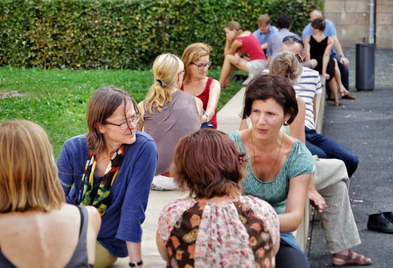 KI generiert: Das Bild zeigt eine Gruppe von Menschen, die im Freien auf einer Bank und auf dem Boden sitzen und sich in kleinen Gruppen unterhalten. Der Hauptinhalt des Bildes ist eine gesellige Zusammenkunft oder ein Treffen in einem Park oder einer ähnlichen Umgebung.