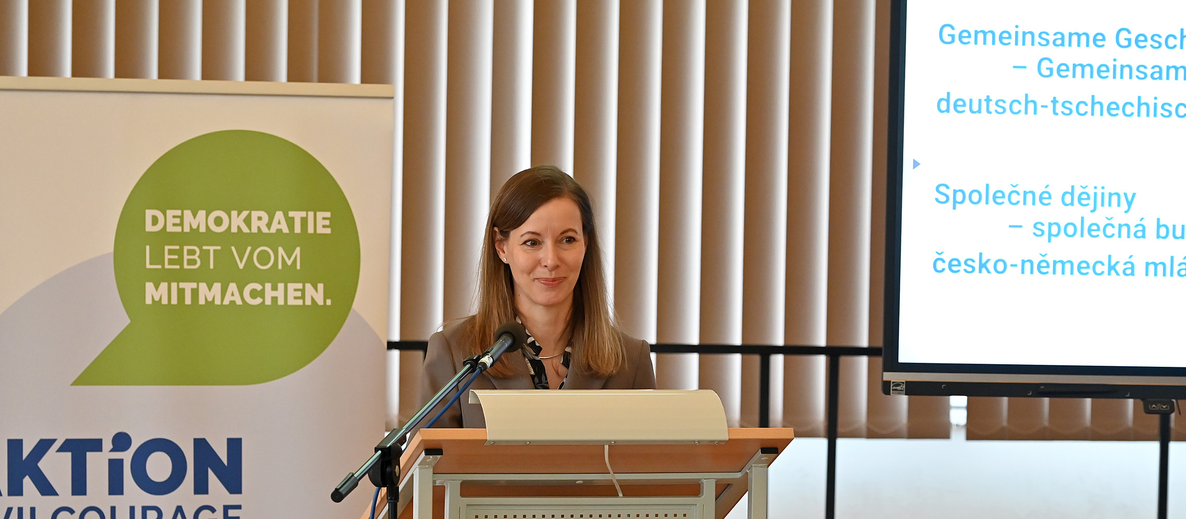 KI generiert: Das Bild zeigt eine Frau, die an einem Pult steht und eine Rede hält. Im Hintergrund sind ein Banner mit der Aufschrift "DEMOKRATIE LEBT VOM MITMACHEN." und eine Projektionsleinwand mit Text in deutscher und tschechischer Sprache zu sehen.