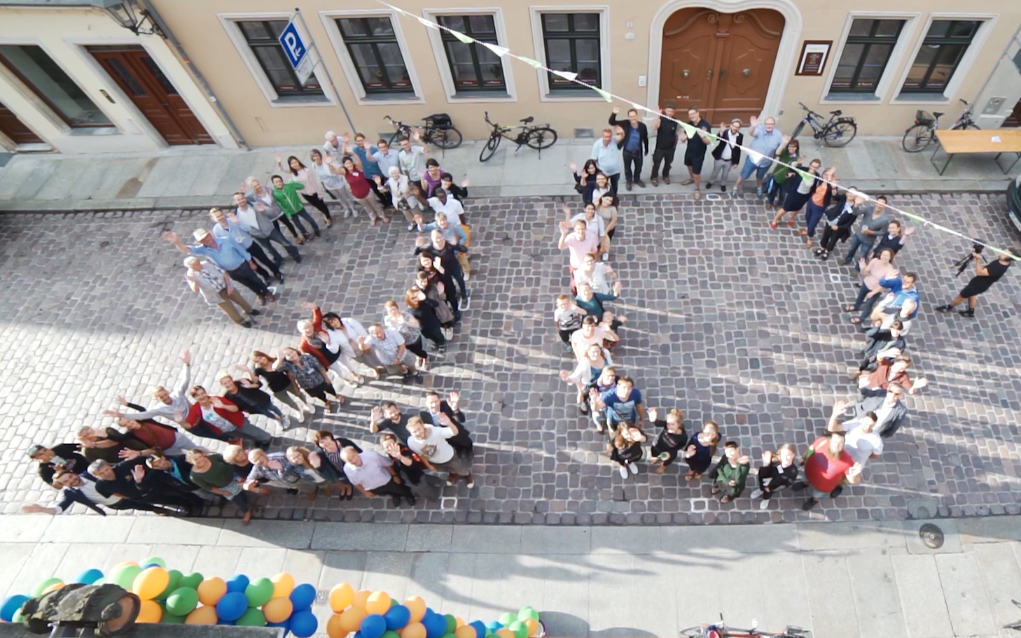 KI generiert: Das Bild zeigt eine große Gruppe von Menschen, die sich auf einem gepflasterten Platz zu einer Formation aufgestellt haben, um ein Wort oder eine Form zu bilden. Im Vordergrund sind bunte Ballons zu sehen, die an einer Absperrung befestigt sind.
