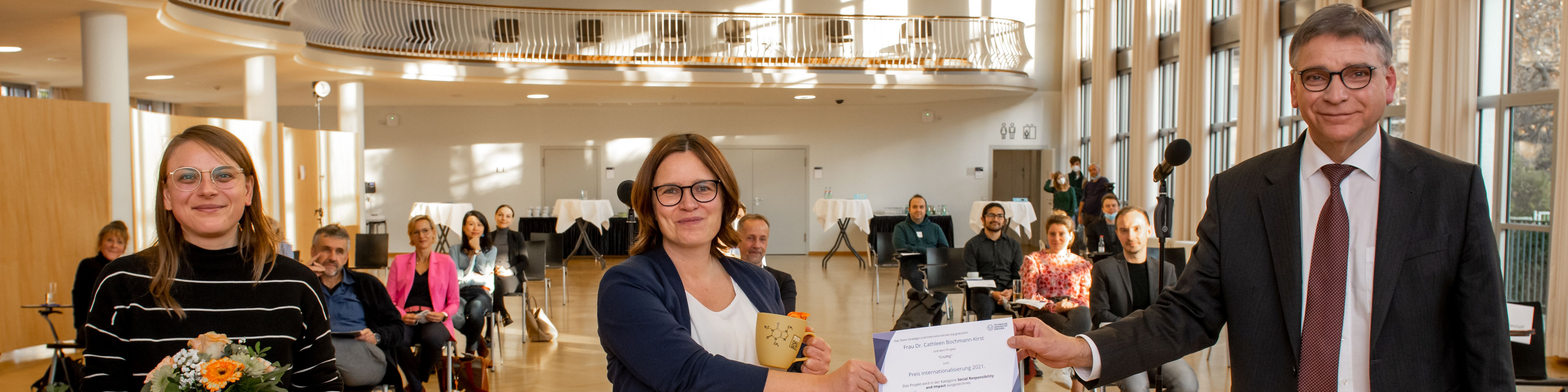 KI generiert: Das Bild zeigt eine offizielle Zeremonie in einem hellen, geräumigen Raum, bei der eine Frau von einem Mann in Anzug eine Urkunde und eine kleine Trophäe erhält, während eine weitere Frau mit Blumen im Arm daneben steht. Im Hintergrund sind mehrere Personen als Publikum anwesend.