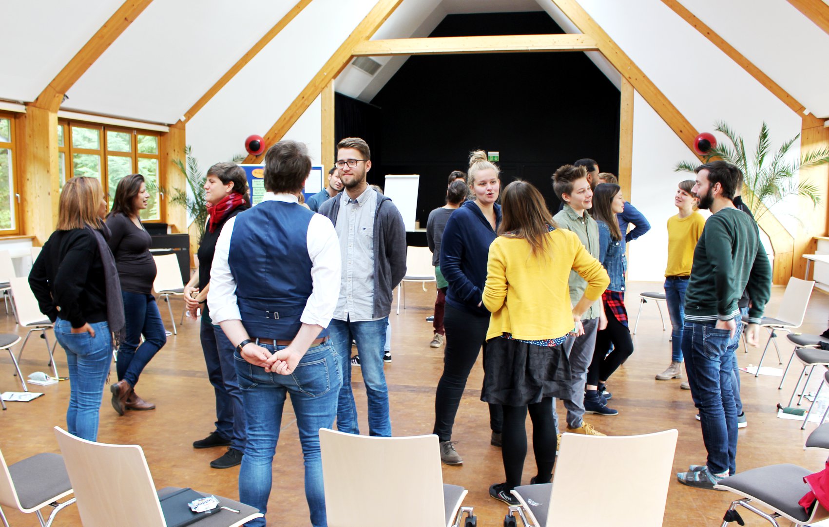 KI generiert: Das Bild zeigt eine Gruppe von Menschen, die in einem hellen, geräumigen Raum mit Holzvertäfelung und großen Fenstern stehen und sich unterhalten. Der Hauptinhalt des Bildes ist eine informelle Zusammenkunft oder ein Workshop in einem Seminarraum.