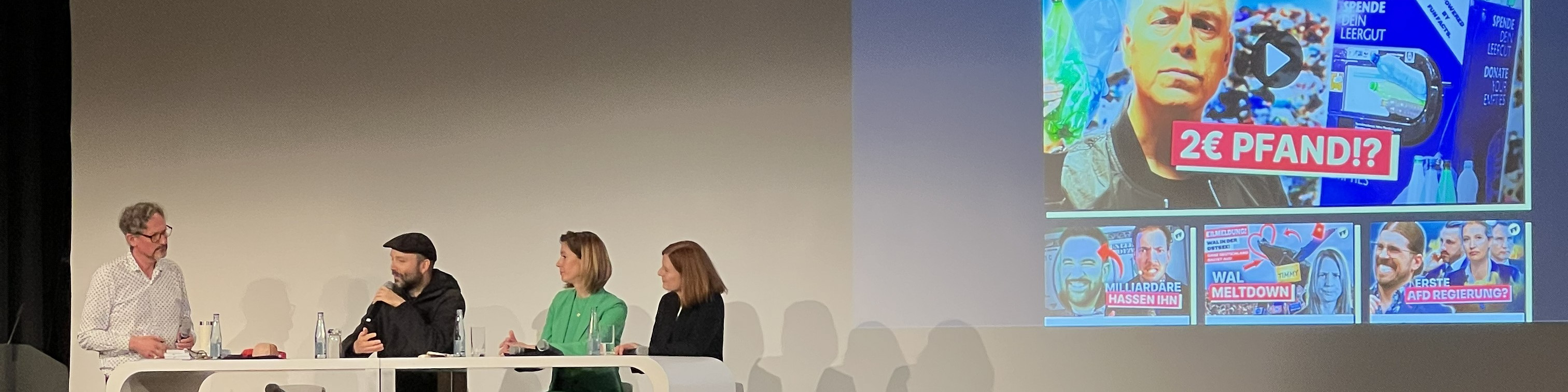 KI generiert: Das Bild zeigt eine Podiumsdiskussion mit vier Personen auf einer Bühne, während im Hintergrund ein großer Bildschirm mit einer Präsentation und der Frage "2€ PFAND?" zu sehen ist. Der Hauptinhalt des Bildes ist die Diskussion, die sich möglicherweise auf das Thema Pfand auf Getränkeverpackungen bezieht.