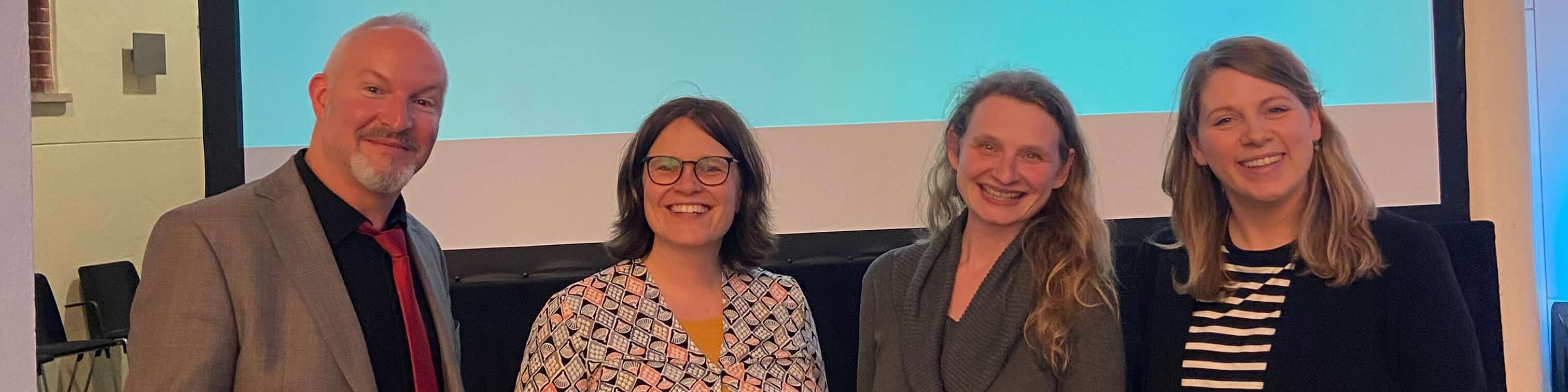 KI generiert: Das Bild zeigt vier lächelnde Personen, die nebeneinander vor einem Projektor oder Bildschirm mit blau-weißen Hintergrund stehen. Der Hauptinhalt des Bildes ist eine Gruppe von Personen, die möglicherweise an einer Präsentation oder einem Meeting teilnehmen.