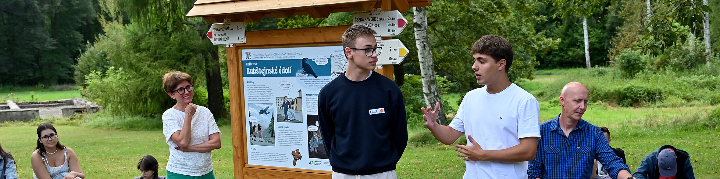 KI generiert: Das Bild zeigt eine Gruppe von Personen im Freien, wobei zwei junge Männer im Vordergrund stehen und sich unterhalten, während eine Informations-tafel und Wegweiser im Hintergrund zu sehen sind. Der Hauptinhalt des Bildes scheint eine Diskussion oder Führung in einer natürlichen Umgebung, möglicherweise einem Park oder Wald, zu sein.