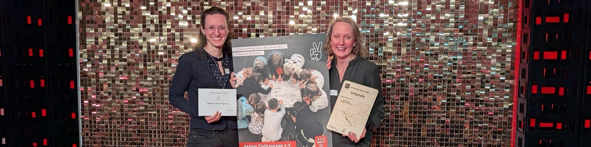 KI generiert: Das Bild zeigt zwei lächelnde Frauen, die jeweils eine Urkunde und ein Buch in den Händen halten, vor einer glänzenden, mosaikartigen Wand. Der Hauptinhalt des Bildes ist die Verleihung oder Anerkennung, die durch die Urkunden symbolisiert wird.