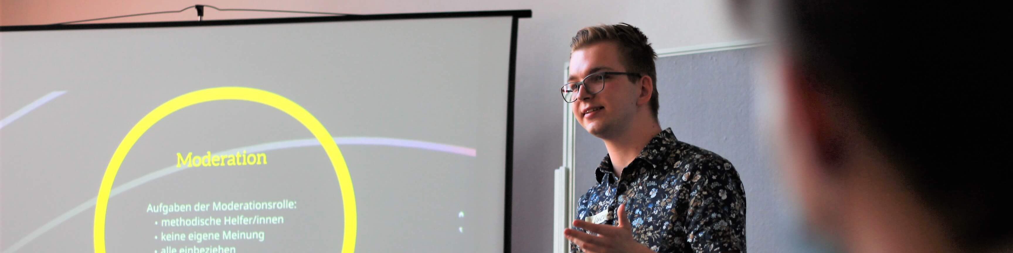 KI generiert: Das Bild zeigt einen Mann, der vor einem Projektor steht und eine Präsentation hält. Der Hauptinhalt der Präsentation scheint das Thema "Moderation" zu sein, wobei spezifische Punkte wie die Aufgaben der Moderationsrolle und methodische Hilfsmittel erwähnt werden.