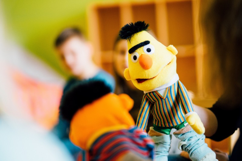 KI generiert: Das Bild zeigt eine Nahaufnahme einer Puppe, die Bert aus der Sesamstraße darstellt. Im Hintergrund sind unscharf zwei Personen zu erkennen.