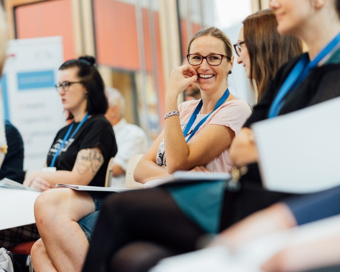 KI generiert: Das Bild zeigt eine Gruppe von Menschen in einem Innenraum, wobei eine lächelnde Frau mit Brille im Vordergrund zu sehen ist. Der Hauptinhalt des Bildes ist eine gesellige Versammlung oder ein Treffen von Personen.