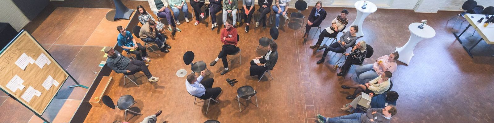 KI generiert: Das Bild zeigt eine Gruppe von Menschen, die in einem großen, hellen Raum mit Holzboden in einem Kreis sitzen und sich unterhalten. Im Vordergrund sind einige Personen, die auf Stühlen sitzen, während andere auf dem Boden Platz genommen haben, was auf eine informelle Diskussion oder ein Meeting hindeutet.