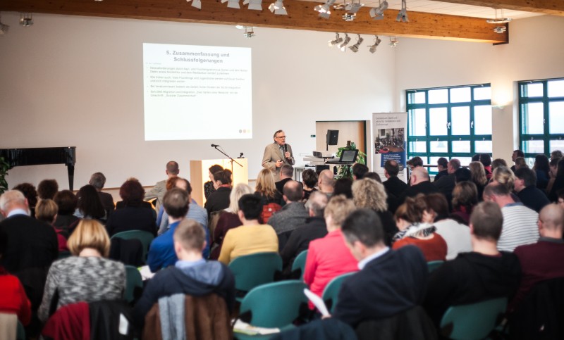 KI generiert: Das Bild zeigt eine Versammlung oder Konferenz in einem hellen Raum mit vielen Teilnehmern, die einem Redner zuhören, der an einem Pult steht. Im Hintergrund ist eine Projektionsleinwand mit einer Präsentation zu sehen, die den Titel "5. Zusammenfassung und Schlussfolgerungen" trägt.