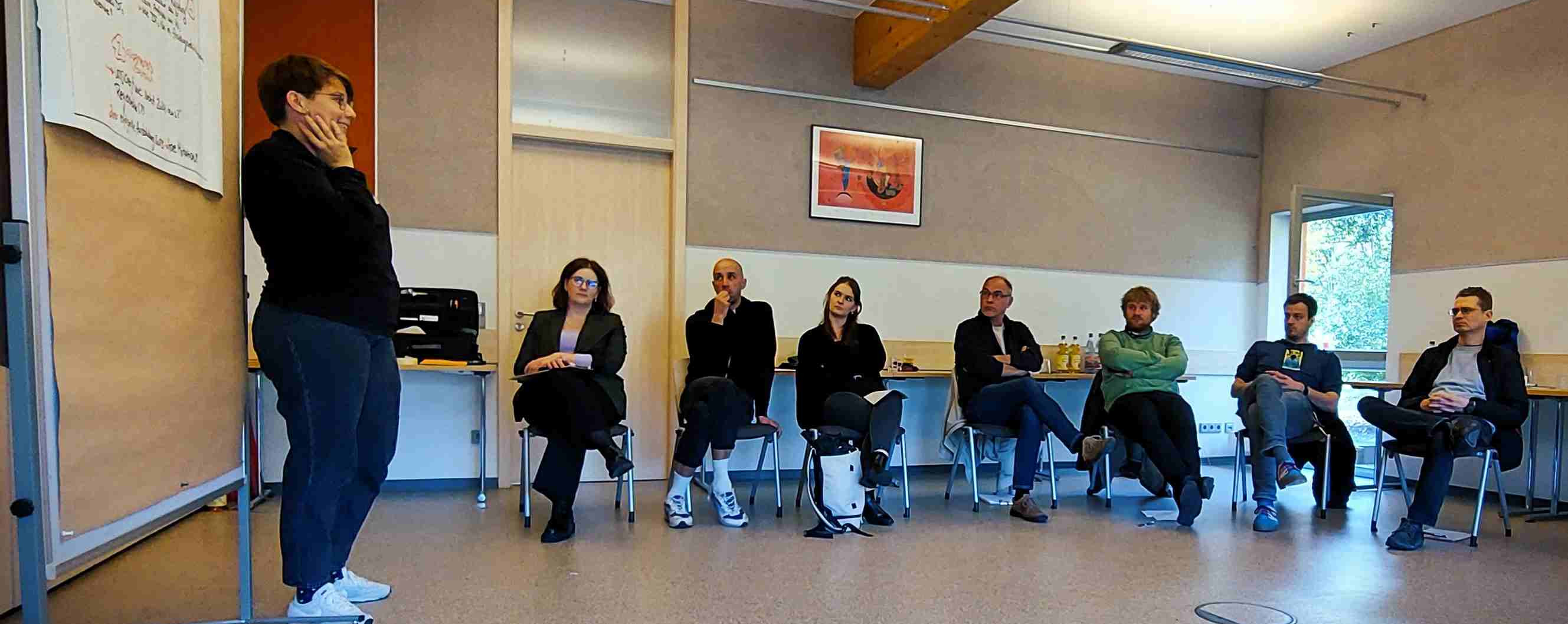 KI generiert: Das Bild zeigt eine Gruppe von Personen in einem Seminarraum, wobei eine Person vorne steht und zu sprechen scheint, während die anderen auf Stühlen sitzen und zuhören. Der Hauptinhalt des Bildes ist eine scheinbare Diskussion oder ein Workshop in einem Klassenzimmer oder Besprechungsraum.