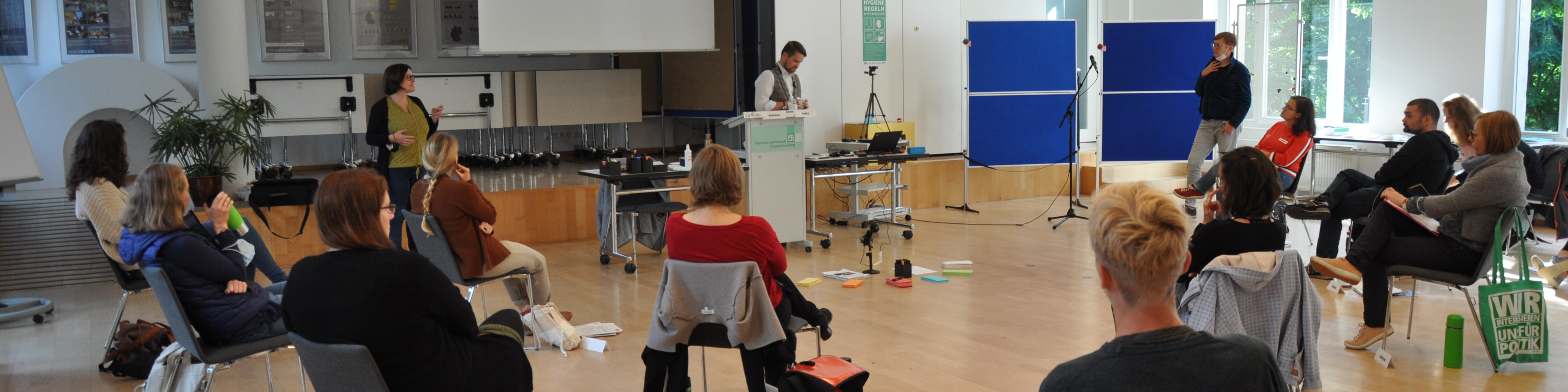 KI generiert: Das Bild zeigt eine Gruppe von Personen in einem hellen Raum, die an einer Präsentation oder einem Workshop teilnehmen. Im Vordergrund sitzen die Zuhörer, während im Hintergrund zwei Personen vor einem Podium und einer Leinwand stehen und sprechen.