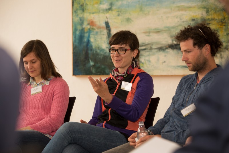 KI generiert: Das Bild zeigt drei Personen, die in einer informellen Sitzung oder Diskussion zusammenkommen, wobei die Person in der Mitte gerade spricht oder eine Geste macht. Im Hintergrund hängt ein abstraktes Gemälde an der Wand.