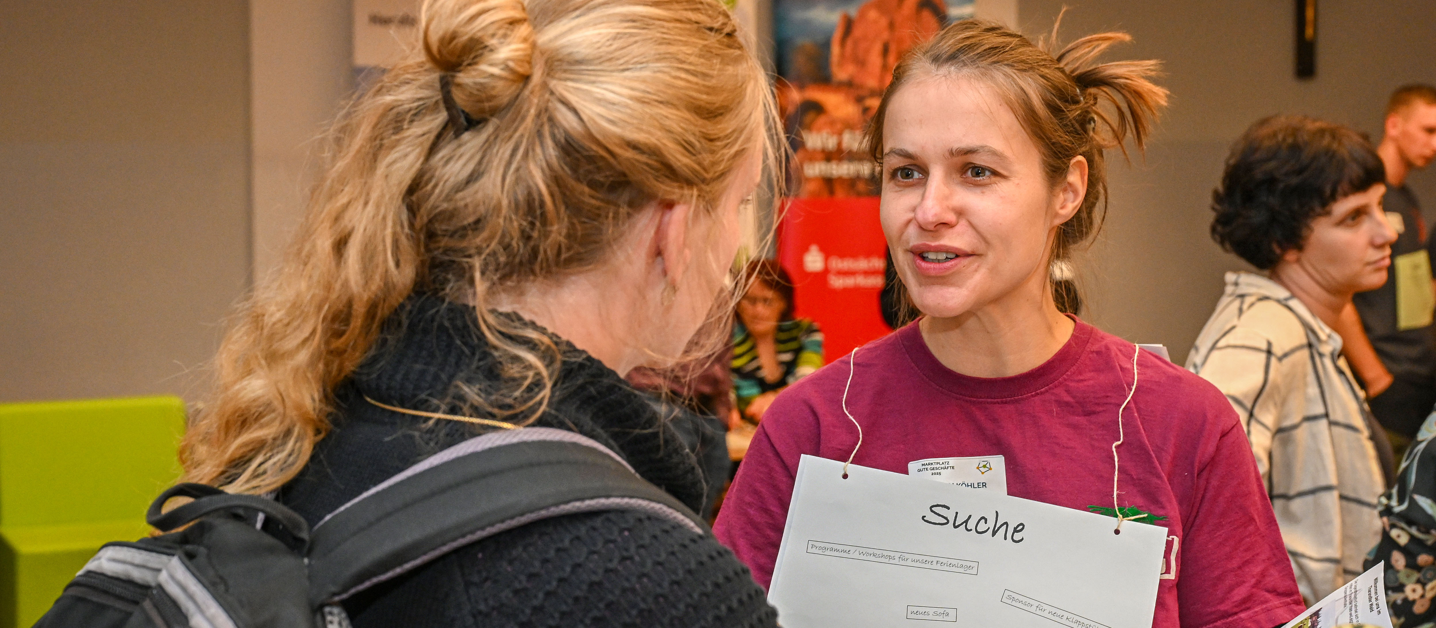 KI generiert: Das Bild zeigt zwei Frauen in einem Gespräch auf einer Veranstaltung, wobei eine der Frauen ein Namensschild mit der Aufschrift "Suche" trägt. Im Hintergrund sind weitere Personen und ein Banner zu sehen, was auf eine organisierte Veranstaltung oder Messe hindeutet.