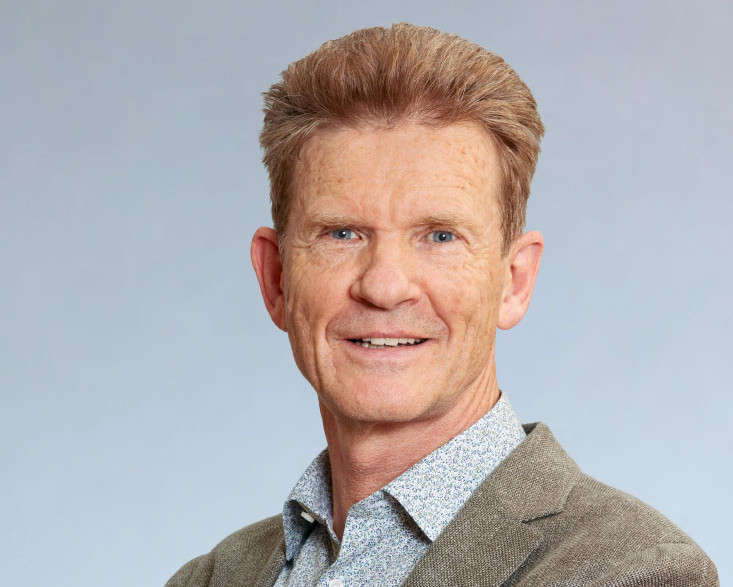 KI generiert: Das Bild zeigt einen lächelnden Mann mit kurzen, rötlichen Haaren, der eine Brille trägt und ein helles, gemustertes Hemd unter einem beigen Sakko anhat. Der Hauptinhalt des Bildes ist das Porträt des Mannes vor einem neutralen, hellen Hintergrund.