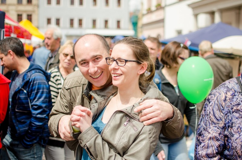 KI generiert: Das Bild zeigt ein fröhliches Paar, das sich auf einer belebten Veranstaltung oder einem Fest umarmt und lachend in die Kamera blickt. Im Hintergrund sind weitere Menschen, bunte Stände und ein grüner Luftballon zu sehen.
