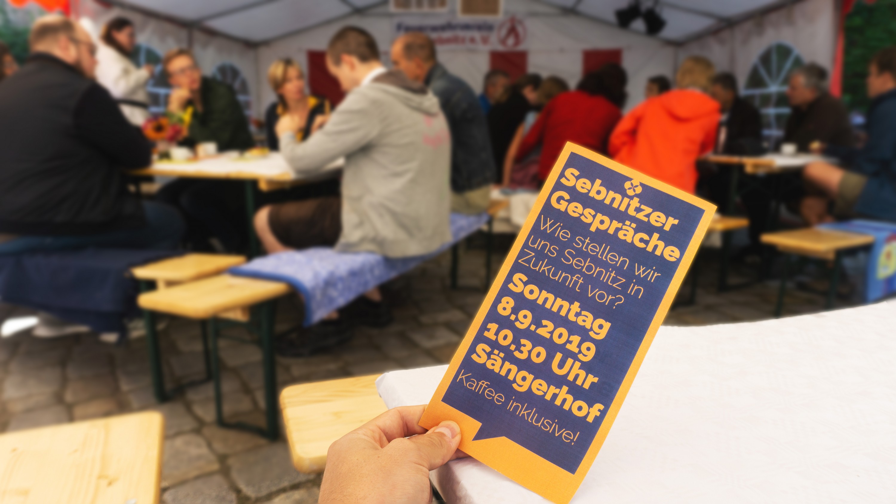KI generiert: Das Bild zeigt eine Person, die eine Einladungskarte für ein "Sebnitzer Gespräch" hält, das am Sonntag, den 8.9.2019 um 10:30 Uhr im Sängerhof stattfindet, inklusive Kaffee. Im Hintergrund sind Menschen in einem Zelt, die sich unterhalten und an Tischen sitzen.