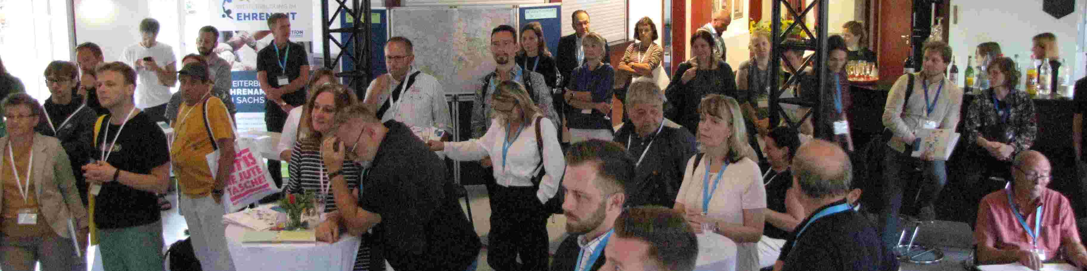 KI generiert: Das Bild zeigt eine Gruppe von Menschen, die sich in einem Innenraum versammelt haben, möglicherweise bei einer Konferenz oder einem Event. Die Teilnehmer stehen und unterhalten sich, einige tragen Namensschilder und halten Getränke in den Händen.