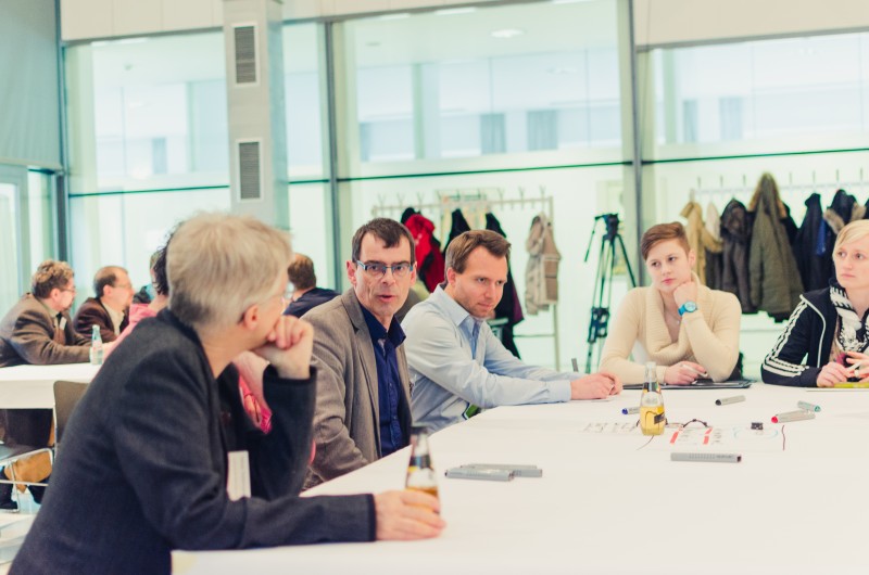 KI generiert: Das Bild zeigt eine Gruppe von Personen, die an einem langen, weißen Tisch in einem hellen Raum mit großen Fenstern sitzen und sich unterhalten. Der Hauptinhalt des Bildes ist eine Besprechung oder ein Meeting in einem modernen, gut beleuchteten Umfeld.