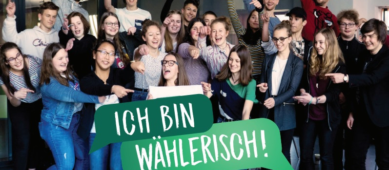 KI generiert: Das Bild zeigt eine Gruppe junger Menschen, die gemeinsam ein Schild mit der Aufschrift "ICH BIN WÄHLERISCH!" halten und dabei fröhlich in die Kamera lächeln. Der Hauptinhalt des Bildes ist die Betonung der Bedeutung von Wahlberechtigung und politischer Teilhabe.