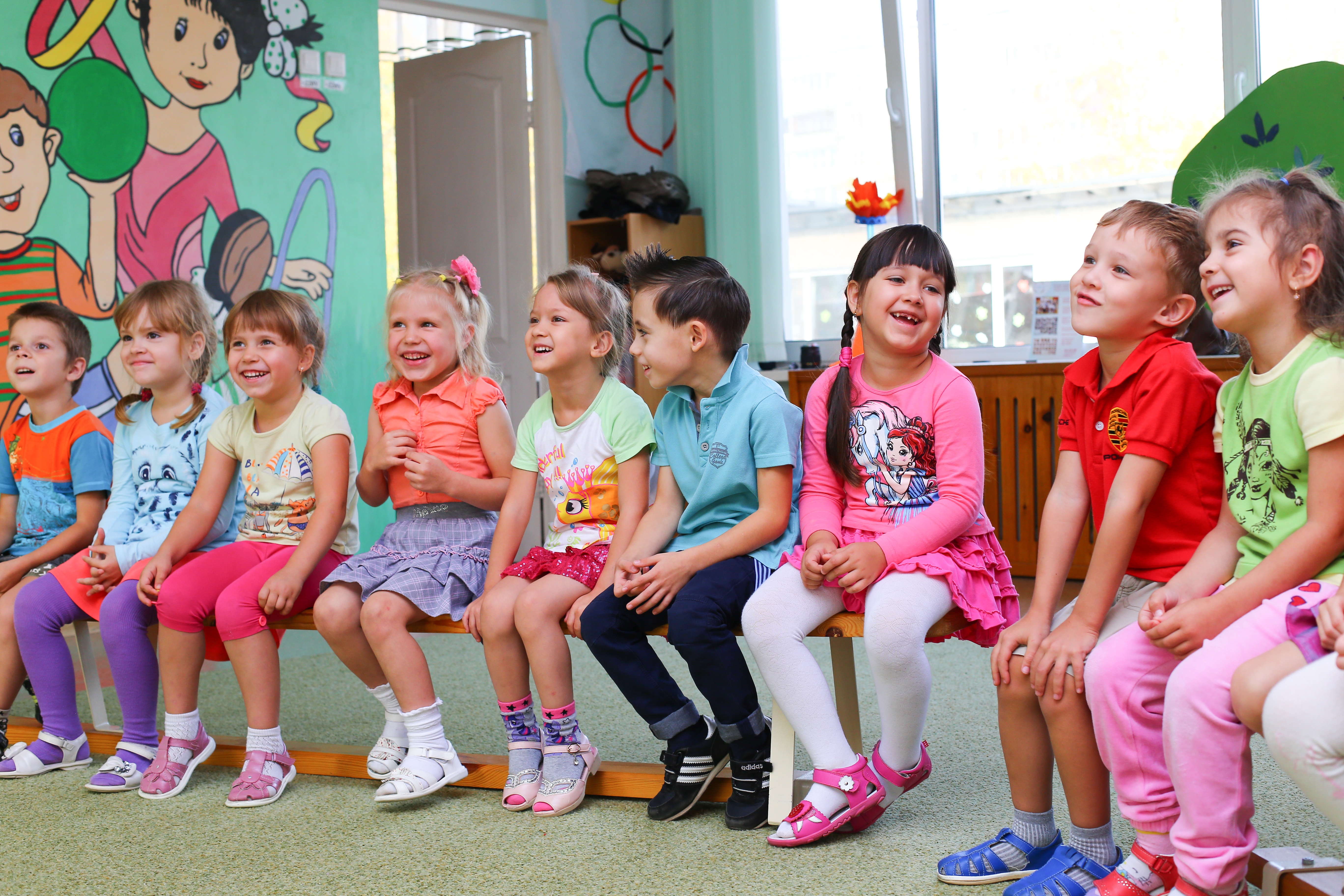 KI generiert: Das Bild zeigt eine Gruppe von Kindern, die auf einer Bank in einem farbenfrohen Raum sitzen und sich unterhalten. Der Hauptinhalt des Bildes ist eine fröhliche und lebendige Kindergruppe in einer scheinbar kindgerechten Umgebung, möglicherweise einem Klassenzimmer oder Spielzimmer.