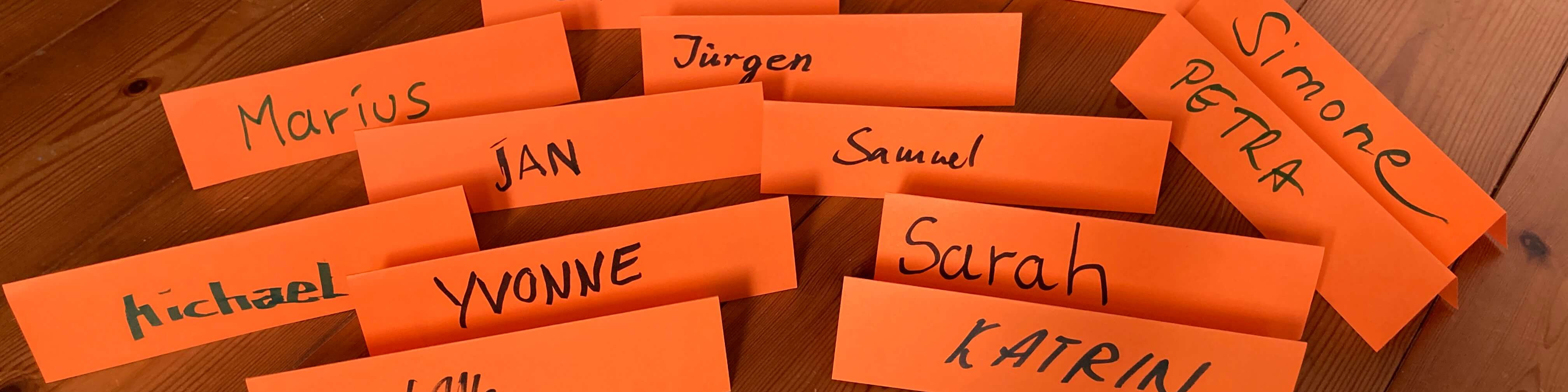KI generiert: Das Bild zeigt mehrere orangefarbene Zettel auf einer Holzfläche, auf denen verschiedene Namen handschriftlich notiert sind. Der Hauptinhalt des Bildes sind diese beschrifteten Zettel mit Namen wie Marius, Jürgen, Samuel, Simone, Sarah, Katrin und anderen.