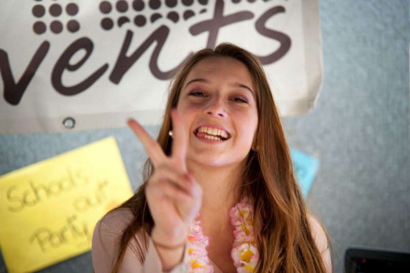 KI generiert: Das Bild zeigt eine lächelnde junge Frau mit einer Blumenkette um den Hals, die den Peace-Zeichen mit den Fingern formt. Im Hintergrund sind ein Banner mit der Aufschrift "Events" und ein Schild mit "School Party" zu sehen.
