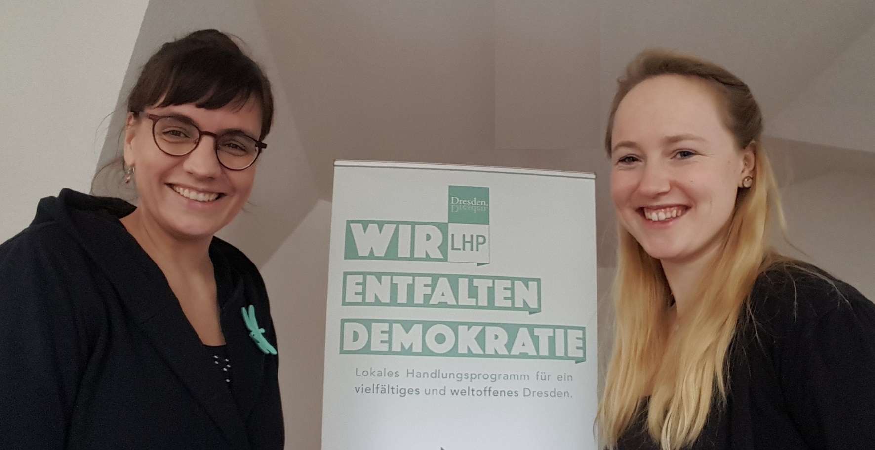 KI generiert: Das Bild zeigt zwei lächelnde Frauen, die neben einem Plakat mit der Aufschrift "Wir entfalten Demokratie" stehen. Das Plakat wirbt für ein lokales Handlungsprogramm für ein vielfältiges und weltoffenes Dresden.